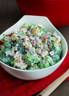 a white bowl filled with broccoli salad on top of a red cloth next to a wooden spoon