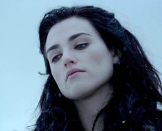 a woman with long black hair and blue eyes looks off into the distance while standing in snow