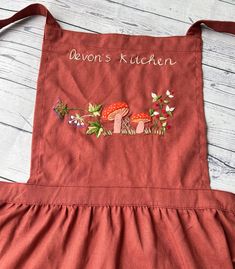 a red apron with embroidered mushrooms and flowers on the front that says baron's kitchen