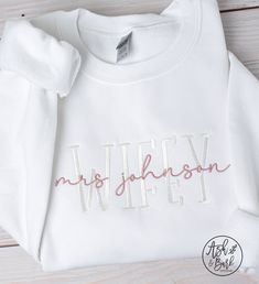 a white shirt with the word wife embroidered on it, sitting on a wooden surface