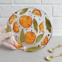 a hand holding a glass plate with oranges on it next to gold spoons