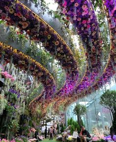 people are sitting on benches in the middle of an indoor garden area with flowers hanging from the ceiling