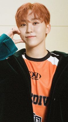 a young man with red hair wearing an orange and black shirt, standing in front of a white wall