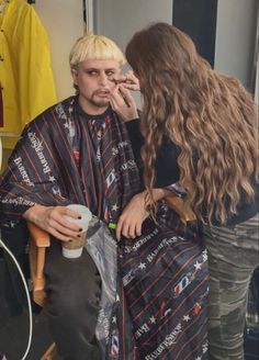 a woman sitting next to a man in a barbershop with blonde hair and makeup