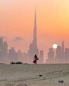 a woman walking on the beach in front of a large city with tall buildings at sunset