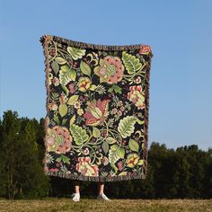 a woman is standing in front of a blanket with flowers on it and trees behind her