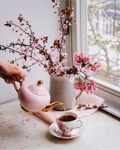 a person pouring tea into a cup on a table next to a vase with pink flowers