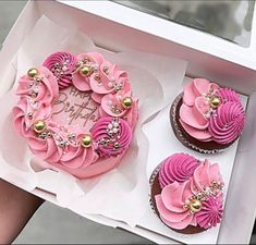 two cupcakes with pink frosting and gold decorations are in a white box