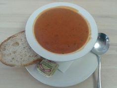 a white plate topped with a bowl of soup next to a slice of bread and a spoon