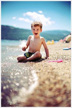 a young boy sitting on the beach with his feet in the water and smiling at the camera