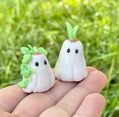 two small white ghost figurines with green leaves on their heads are held in the palm of someone's hand