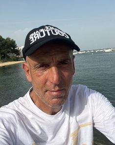 a man wearing a white shirt and black hat sitting in front of the water with his hands on his hips