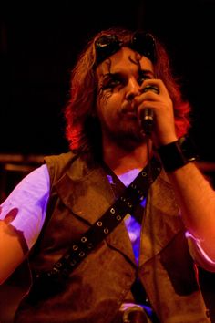 a man with makeup on his face holding a microphone to his ear and wearing a vest