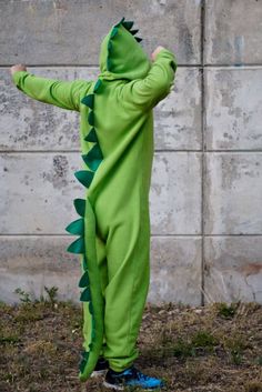 a little boy in a green dinosaur costume standing on the grass with his arms outstretched