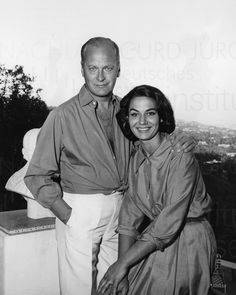 an older man and woman standing next to each other in front of a cityscape