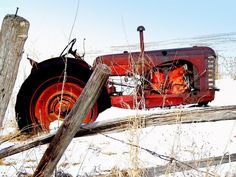 an old red tractor sitting on top of snow covered ground next to a wooden fence