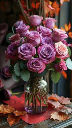 a vase filled with purple roses sitting on top of a table next to autumn leaves