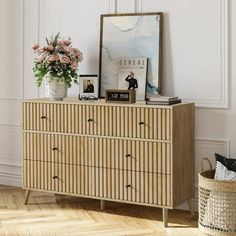 a wooden dresser with flowers and pictures on it in a white walled room next to a wicker basket