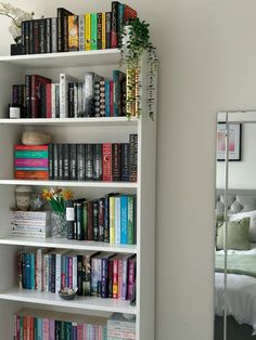 a bookshelf filled with lots of books next to a white bed and mirror