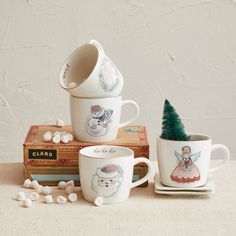 three mugs with designs on them sitting next to some books and a small christmas tree