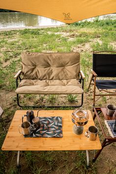 two chairs and a coffee table in the grass near a body of water with an umbrella over them