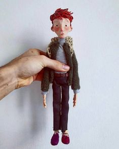 a hand holding a small doll in front of a white wall with a red hair