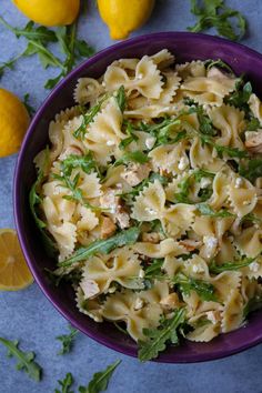 a purple bowl filled with pasta and spinach next to lemons on a blue surface