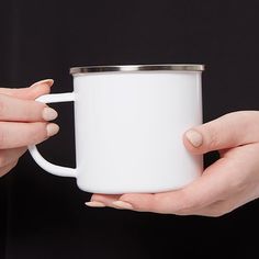 two hands holding a white coffee mug against a black background with the lid off and one hand on the cup