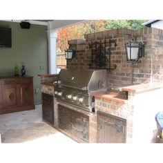 an outdoor kitchen with grill and television mounted on the brick wall, along with wooden cabinets