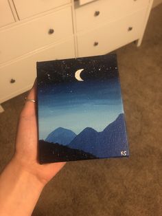 a hand holding up a small piece of art with mountains and the moon in the sky