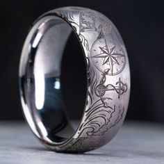 "Celebrate the maritime spirit of your high-seas adventures with this exquisite 8mm wide domed tungsten pirate-themed wedding band. Adorned with iconic nautical symbols including a menacing monsters of the deep, a weathered ship's wheel, a gleaming anchor, and a treasure chest overflowing with doubloons, each meticulously laser etched to create a lasting tribute to your seafaring memories. Our dedication to precision engraving ensures a flawless and sophisticated finish for this ring, featuring wire wheel polishing for a subtle and understated touch. Handcrafted in our humble workshop, using only the finest materials and cutting-edge equipment, each ring is a genuine work of art. Should you desire a custom pirate design that speaks to your unique voyage, do not hesitate to contact us - we' Pirate Wedding Ring, Luxury Round Ring With Compass Design, Vintage Jewelry Men, Pirate Rings, Unique Mens Wedding Rings, Pirate Wedding Theme, Nautical Symbols, Pirate Design, Pirate Wedding