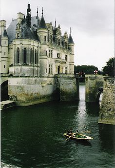 an old castle sitting on the side of a river next to a bridge with a row boat in front of it