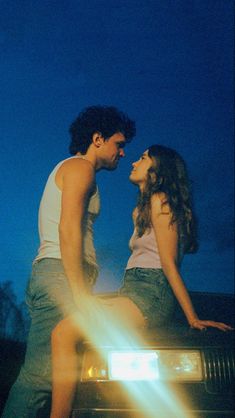 a man and woman sitting on the hood of a car in front of a blue sky