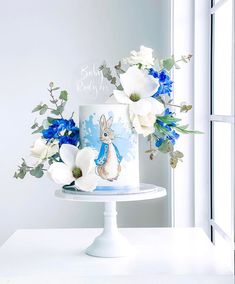 a white cake with blue and white flowers sitting on top of a table next to a window