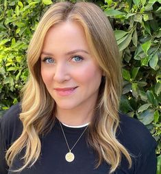 a woman with blonde hair and blue eyes wearing a black shirt in front of bushes