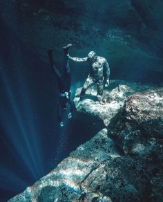 two people in scuba gear standing on rocks near the ocean water with sunlight streaming through them