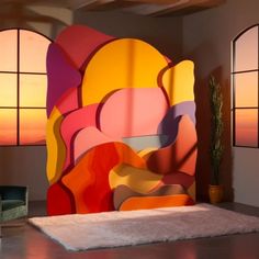 a room with two windows and a large colorful painting on the wall behind an upholstered chair