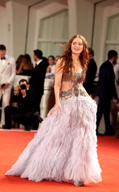 a woman in a long purple dress walking on a red carpet with people behind her