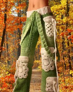 a woman wearing green and white crocheted pants in front of autumn trees with leaves on the ground