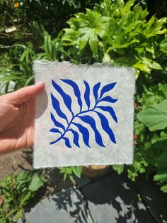 a hand holding up a piece of paper with blue leaves on it in front of some plants