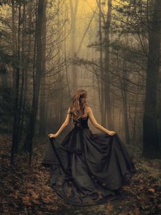 a woman in a black dress is walking through the woods with her back to the camera