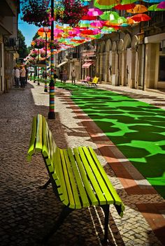 a green bench sitting on the side of a road under colorful umbrellas hanging from it's ceiling