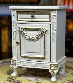 a small white cabinet with gold trimmings on the door and drawer, sitting on a rug