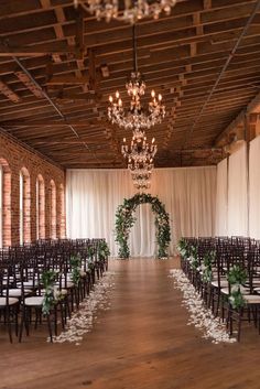 an indoor wedding venue with rows of chairs and chandeliers, decorated with greenery