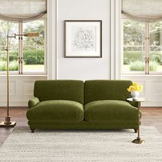 a green couch sitting in front of two windows with curtains on them and a lamp next to it