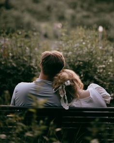 a man and woman sitting on a bench in front of some bushes looking at the sky