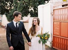 a bride and groom holding hands in front of an orange fence with sunflowers