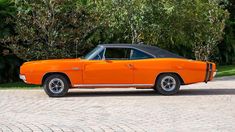 an orange muscle car parked in front of some trees and bushes on a brick road