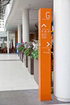 an orange sign on the side of a building near potted plants and people walking by