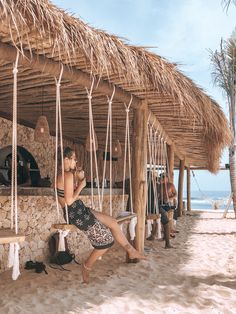 a woman sitting on a bench in front of a hut with thatched roof next to the ocean